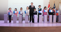 Group of children holding certificates on stage next to an adult, in front of stacked gift boxes.